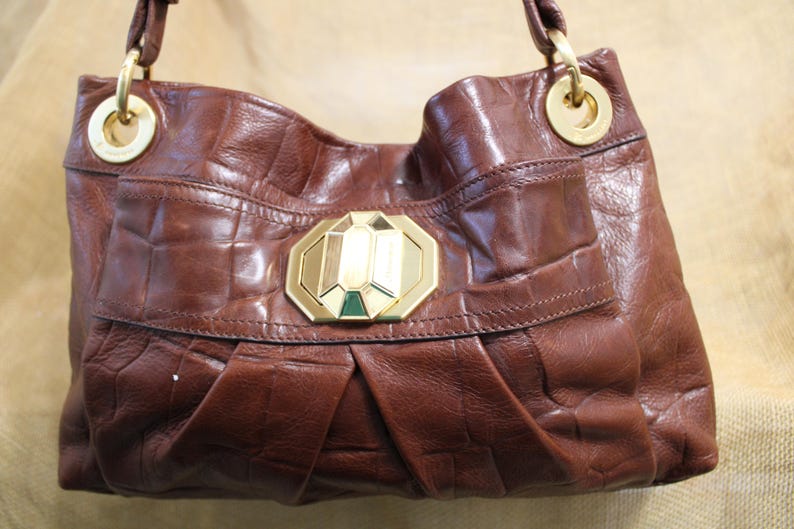 Op de afbeelding: Een bruine leren handtas met een getextureerd, krokodilachtig patroon. De tas heeft een goudkleurige sluiting en hardware, inclusief cirkelvormige ringen voor de handvatten. De voorklep heeft een decoratief goud en groen accent.