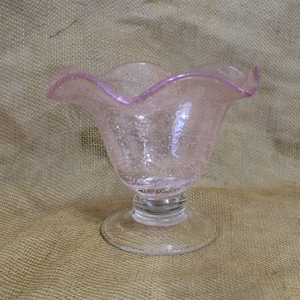 May include: A pink glass bowl with a ruffled rim and a clear glass pedestal base. The bowl has a textured surface with small air bubbles.