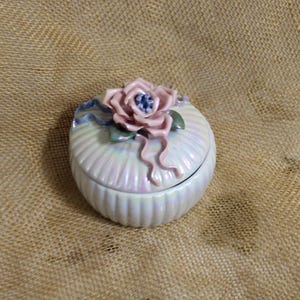 May include: A white ceramic trinket box with a pink flower design on the lid. The box has a ribbed texture and a pearlescent finish.