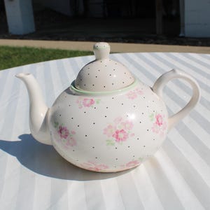 May include: A white teapot with a floral pattern and black polka dots. The teapot has a lid with a green trim and a spout on the side.