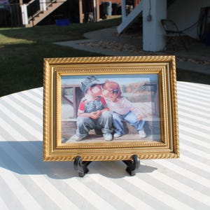 May include: A framed picture with a gold-colored ornate frame. The picture inside depicts two children kissing. The frame is displayed on a white and gray striped surface, supported by a black easel.