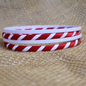 May include: Stack of three bracelets. The top and middle bracelets are white. The bottom bracelet is red and white striped. The bracelets are stacked on top of each other. The background is a tan, textured surface.