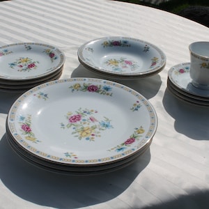 May include: A set of white porcelain dinnerware with floral patterns in shades of pink, yellow, and blue. The set includes a teacup and saucer, a soup bowl, and a dinner plate.