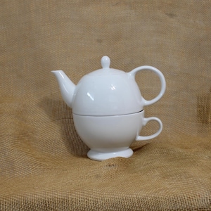 May include: A white ceramic tea set consisting of a teapot stacked directly on top of a matching teacup. The teapot features a rounded body, a curved spout, a handle, and a lid with a small knob. The teacup has a pedestal base and a handle.
