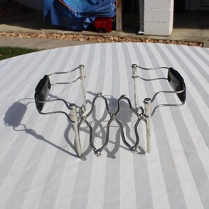 May include: A silver and black expandable wire rack with black plastic handles. The rack is designed to hold a teapot and cups.