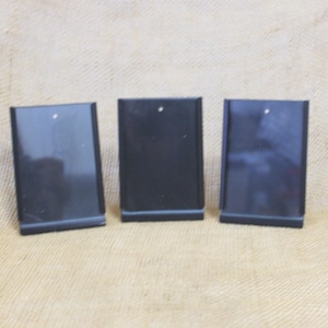 May include: Three black rectangular display stands with a small hole at the top, likely for jewelry. The stands are positioned on a tan, textured surface, suggesting a craft or retail setting. The stands are evenly spaced.