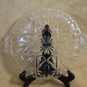 May include: A clear glass oval serving dish with a decorative starburst pattern and two handles. The dish is on a black stand.