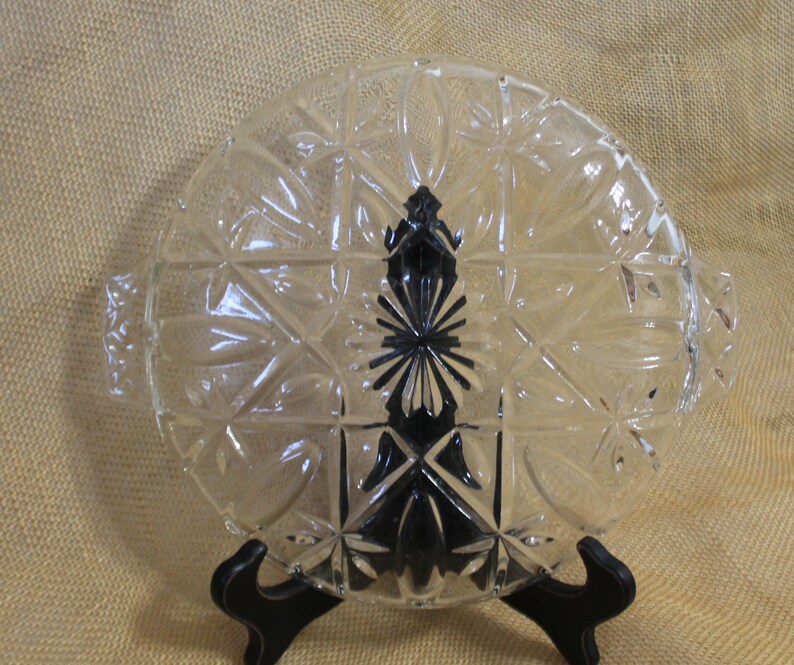 May include: A clear glass oval serving dish with a decorative starburst pattern and two handles. The dish is on a black stand.