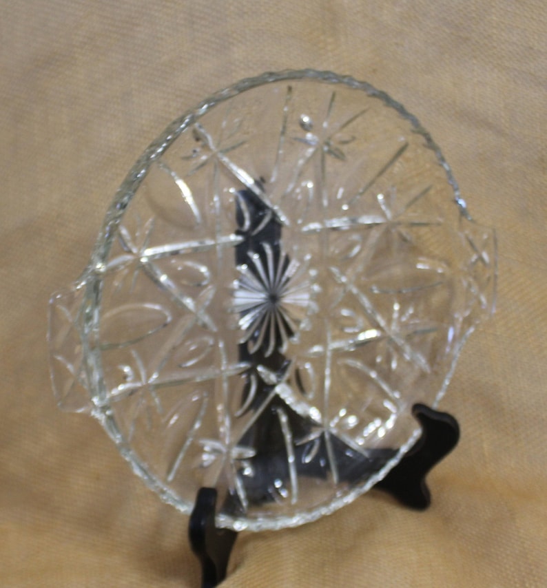 May include: A clear glass serving dish with a starburst pattern and two handles. The dish is on a black stand.