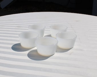 May include: Five small, white, opaque glass bowls are arranged on a white, striped surface. The bowls are cylindrical with a slight taper and rounded bottoms. The lighting creates soft shadows.