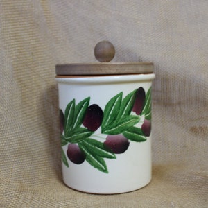 May include: White ceramic jar with a wooden lid and a painted design of green olive branches and purple olives.