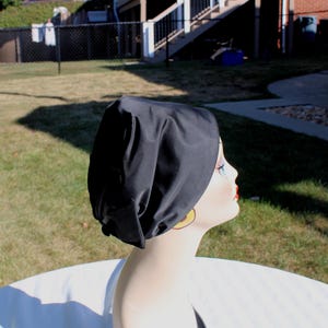 May include: A black head covering, possibly a sleep cap or bonnet, is displayed on a mannequin head. The cap is made of a smooth, dark fabric. The mannequin has blue eyes, red lips, and gold hoop earrings. The background is a grassy yard.