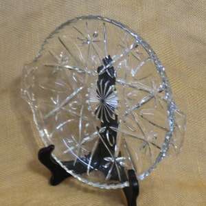 May include: A clear glass serving dish with a scalloped edge and a starburst pattern. The dish is on a black stand.