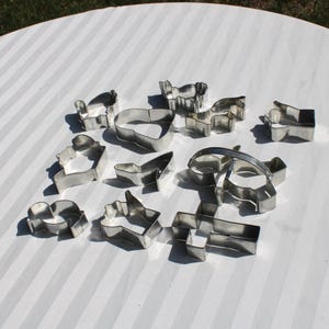 May include: A collection of various stainless steel cookie cutters in different shapes, including animals and geometric forms. The cutters are arranged on a white surface with a striped pattern, ideal for baking.