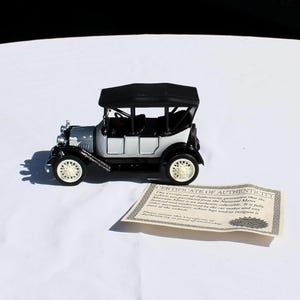May include: A miniature black and silver vintage-style automobile model featuring a black roof, white spoked wheels, and a spare tire mounted on the rear. Beside the vehicle lies a rectangular paper Certificate of Authenticity with printed text.