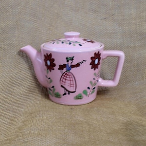 May include: A pink ceramic teapot with a lid, spout, and handle. The teapot is decorated with a floral pattern on the lid and sides, and a painted figure of a woman in a skirt. The teapot is on a tan surface.