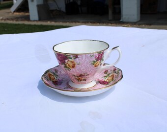 May include: A pink teacup and saucer set with a floral pattern. The teacup and saucer have a gold trim. The teacup has a handle. The floral pattern includes yellow, pink, and purple roses.