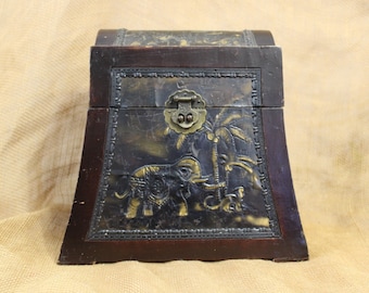May include: A wooden box with a hinged lid and a brass latch. The box is decorated with an embossed design of an elephant and a palm tree. The box is dark brown with a black and gold finish.