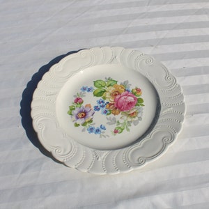 May include: A white ceramic plate with a raised, scalloped edge. The center of the plate features a floral design with pink, yellow, and purple flowers.