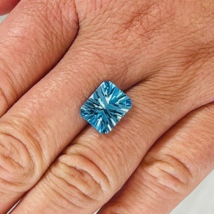 May include: A square-shaped, light blue gemstone with a faceted cut. The gemstone is set in a ring and is being worn on a finger.
