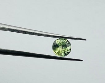May include: A round, green gemstone held by tweezers. The gemstone is a light green color with a slight yellow hue.