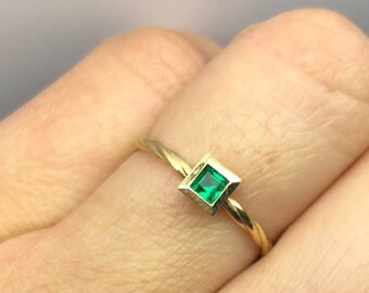 May include: A gold ring featuring a square, emerald-green gemstone. The ring is simple in design, with a thin band and a bezel setting for the gemstone. The ring is shown on a person's finger.