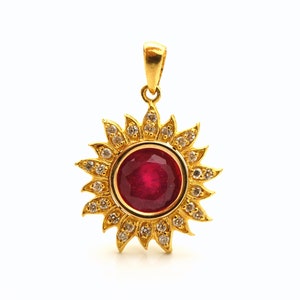 May include: A gold pendant featuring a red ruby gemstone surrounded by a halo of smaller white gemstones, set in a sunburst design.