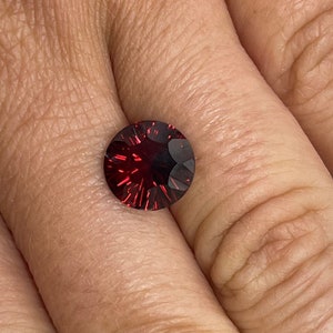 May include: A round, red gemstone with a faceted cut. The gemstone is set in a ring and is being worn on a finger.