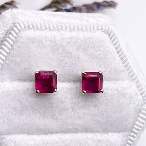 May include: Gold stud earrings, each featuring a square-cut, ruby-red gemstone. The earrings are presented on a white, velvet-lined box, with lavender sprigs in the background. The gemstones have a rich, saturated colour.