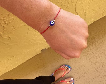 May include: A red string bracelet with a blue evil eye charm. The bracelet is tied around a person's wrist.