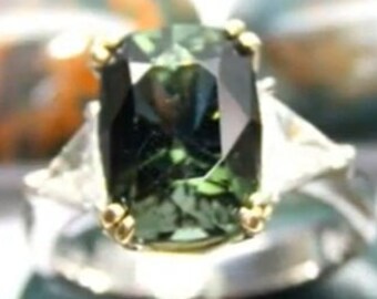 May include: A silver ring with a large, green gemstone surrounded by smaller, clear gemstones. The ring is set on a background of green and brown stones.