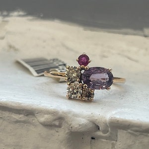 May include: A gold ring with a purple oval gemstone and smaller white and red gemstones.