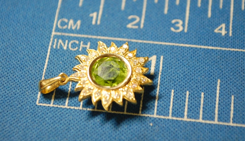 May include: A gold pendant in the shape of a sun with a green gemstone in the center and small white stones around the edge.