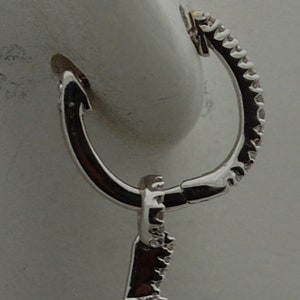 May include: A silver earring with a blue gemstone set in a diamond-shaped halo. The earring has a hoop closure.