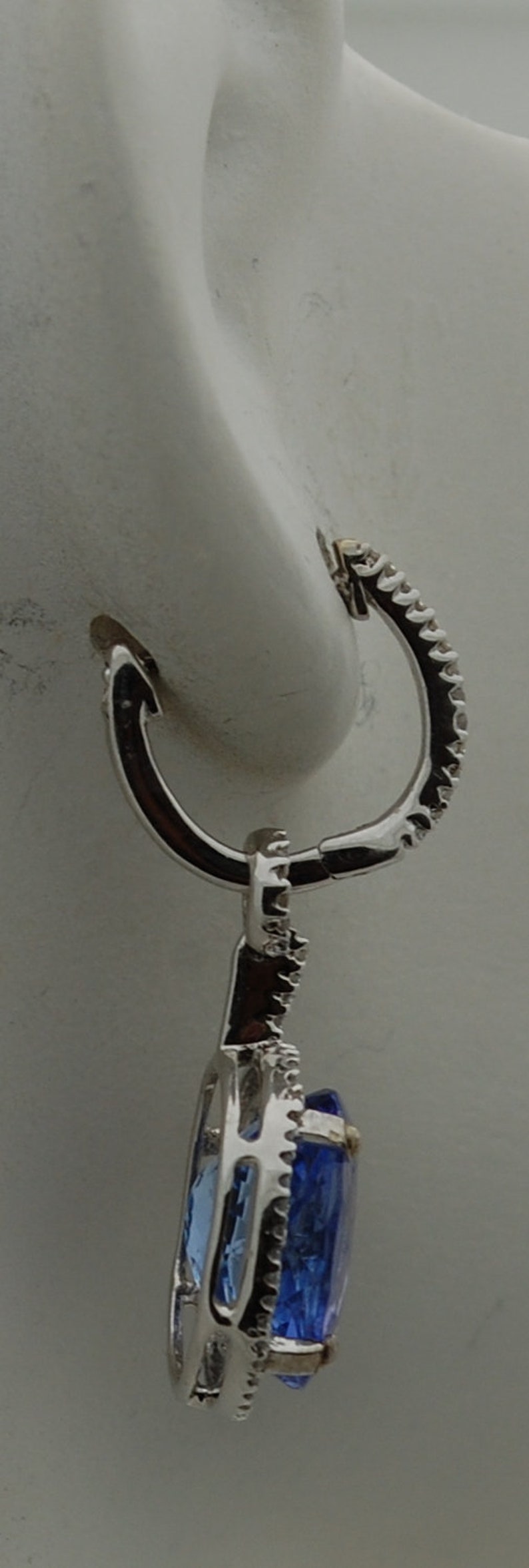 May include: A silver earring with a blue gemstone set in a diamond-shaped halo. The earring has a hoop closure.