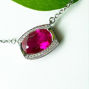May include: A silver chain necklace with a large, oval-shaped, bright pink gemstone pendant set in a silver bezel with a row of small, clear gemstones around the edge.