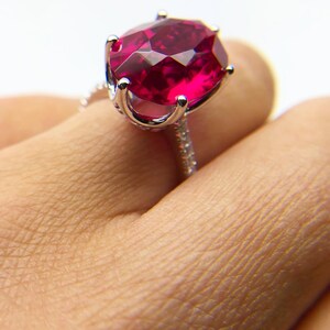 May include: A silver ring with a large, oval-shaped ruby gemstone. The ruby is a deep red color and is set in a prong setting. The ring is on a finger.