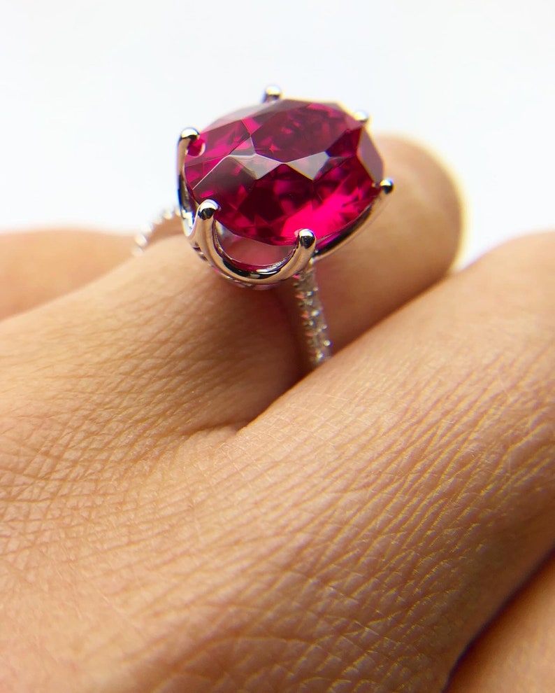 May include: A silver ring with a large, oval-shaped ruby gemstone. The ruby is a deep red color and is set in a prong setting. The ring is on a finger.