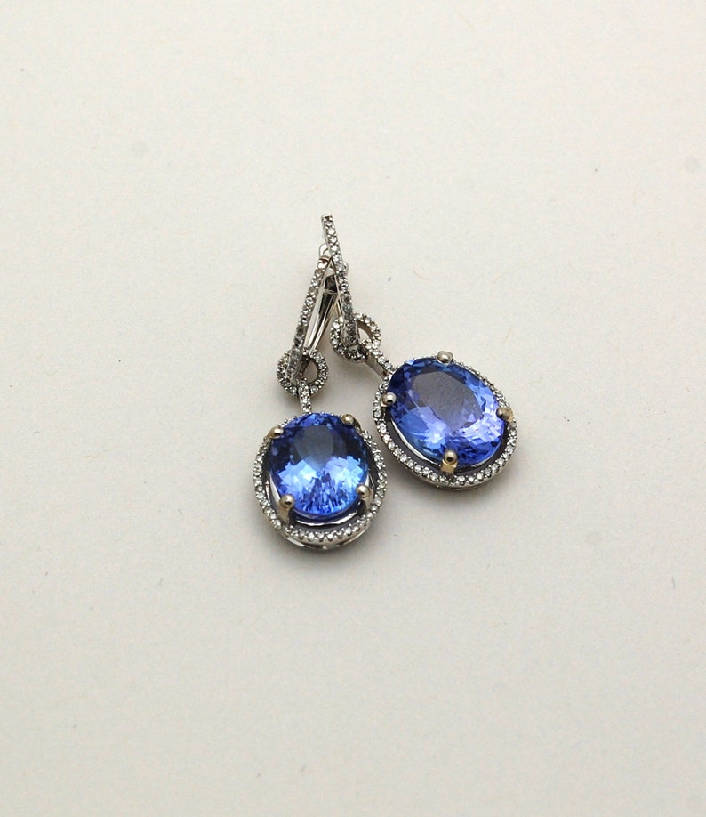 May include: A pair of dangle earrings with oval-shaped blue gemstones surrounded by a halo of small diamonds. The earrings are made of silver and have a drop design.