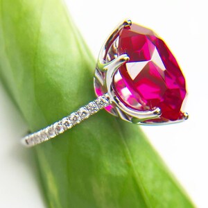 May include: A silver ring with a large, bright red gemstone surrounded by smaller, clear gemstones. The ring is resting on a green leaf.