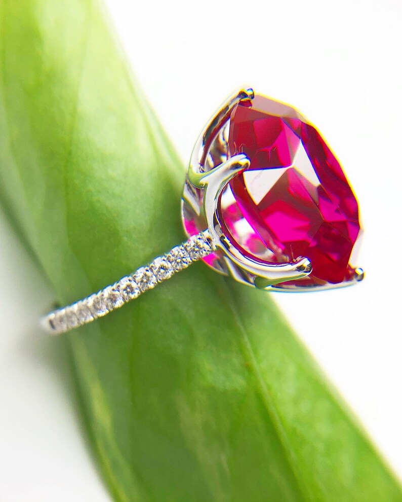 May include: A silver ring with a large, bright red gemstone surrounded by smaller, clear gemstones. The ring is resting on a green leaf.