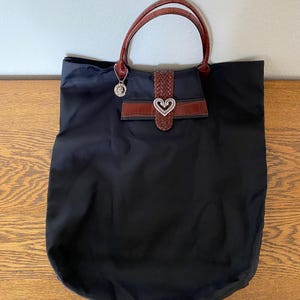 May include: A black tote bag with brown leather handles and a heart-shaped buckle. The bag has a small pocket on the front with a brown leather flap.