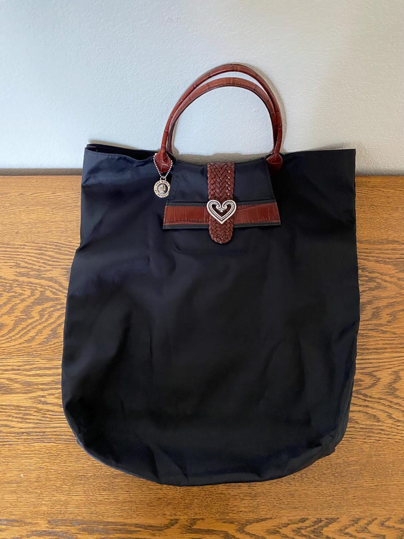 May include: A black tote bag with brown leather handles and a heart-shaped buckle. The bag has a small pocket on the front with a brown leather flap.
