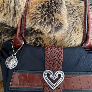 May include: Black handbag with a brown braided strap and a silver heart-shaped buckle. The bag has a brown leather trim and a silver charm that says "One World by Brighton".