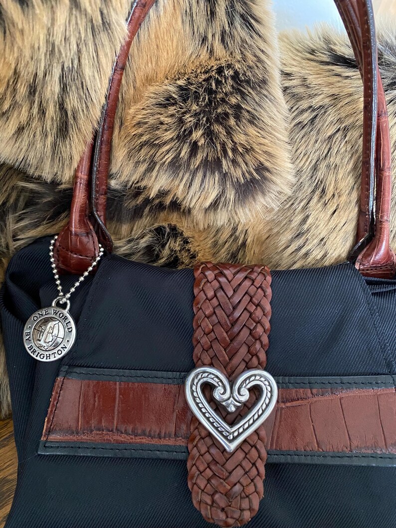 May include: Black handbag with a brown braided strap and a silver heart-shaped buckle. The bag has a brown leather trim and a silver charm that says "One World by Brighton".