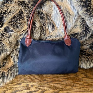 May include: A black nylon handbag with brown leather handles. The bag is resting on a wooden surface.