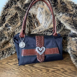 May include: Black nylon handbag with brown leather accents and a silver heart-shaped buckle. The bag has two brown leather handles and a silver charm with the words "One World, One Heart" on it.