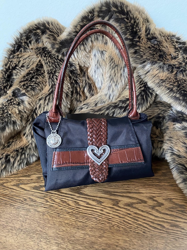 May include: Black nylon handbag with brown leather accents and a silver heart-shaped buckle. The bag has two brown leather handles and a silver charm with the words "One World, One Heart" on it.