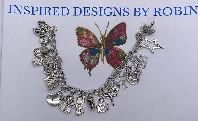 Puede incluir: Pulsera de abalorios plateada con varios dijes, incluyendo un coraz&oacute;n, una cinta, una taza de caf&eacute; y una estrella. El fondo presenta una mariposa colorida y el texto "INSPIRED DESIGNS BY ROBIN".