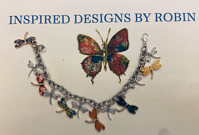 Puede incluir: Un brazalete de plata con cierre de langosta presenta m&uacute;ltiples dijes de lib&eacute;lula en varios colores, incluyendo negro, rojo, amarillo, naranja y verde. Los dijes est&aacute;n detallados con intrincados patrones y dise&ntilde;os. El brazalete est&aacute; adornado con una cadena de plata.